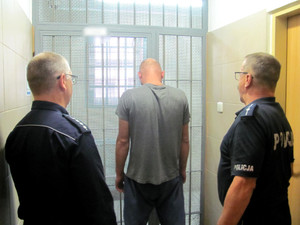 Dwóch policjantów z zatrzymanym mężczyzną stoją przed kratą w pomieszczeniu dla osób zatrzymanych