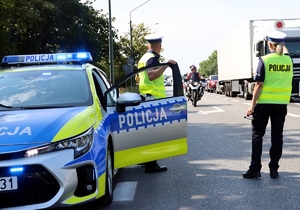 radiowóz i dwoje policjantów na pierwszym planie, motocyklista i pojazdy na drodze