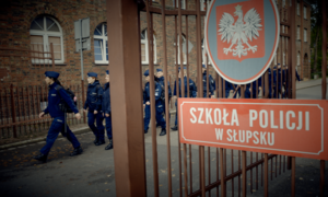 Brama wejściowa na teren Szkoły Policji w Słupsku z tablicą, w tle grupa policjantów
