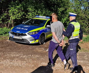policjant drogówki prowadzi kierowcę zatrzymanego po pościgu do stojącego obok radiowozu