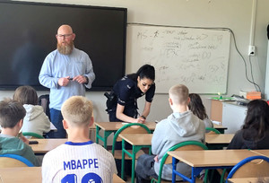 Policjantka i przedstawiciel projektu przed grupa siedzących w ławkach uczniów