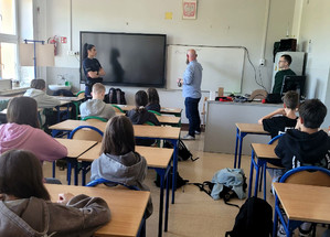 Policjantka i przedstawiciel projektu przed grupą siedzących uczniów w sali