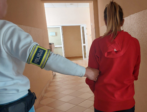 Policjant kryminalny z opaską na ręku prowadzi kobietę korytarzem komendy